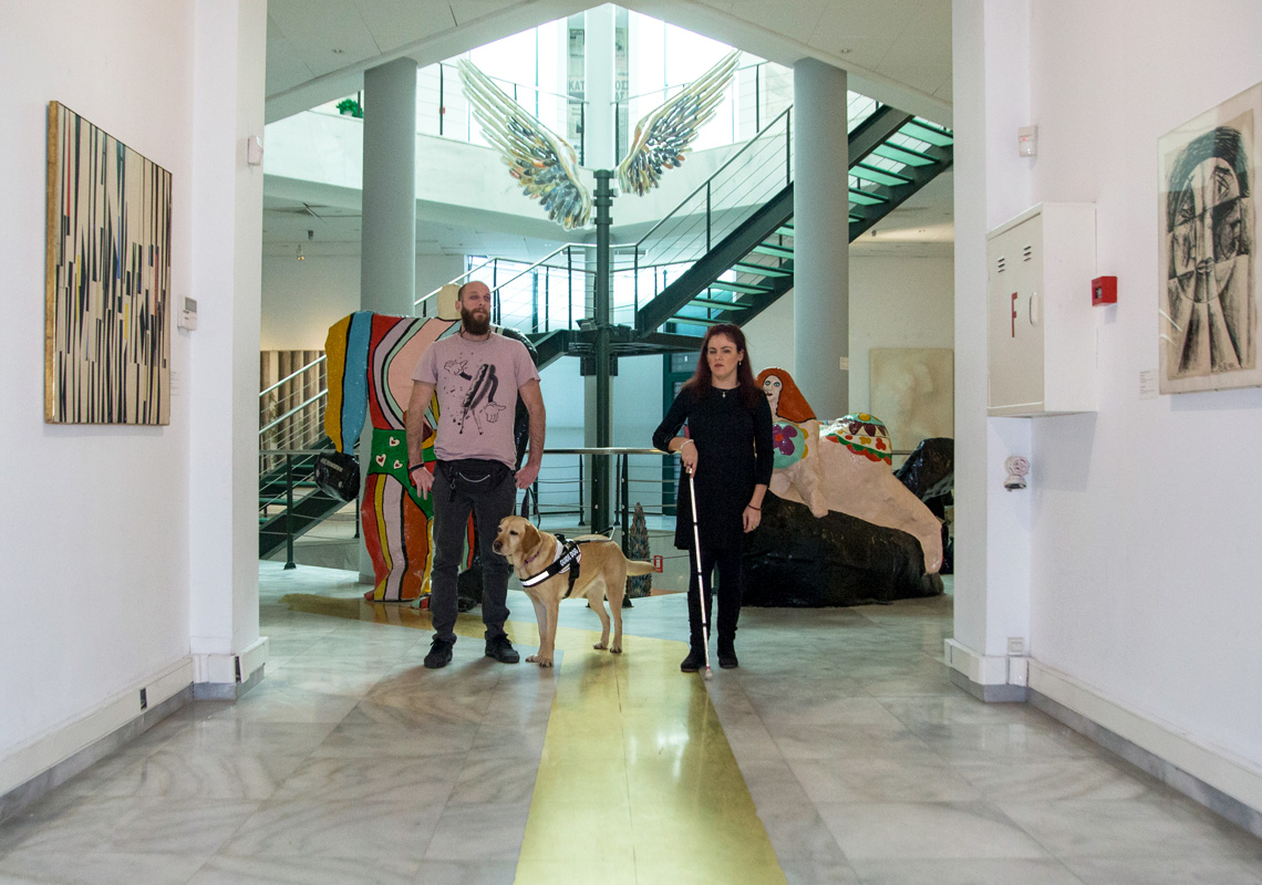 Φωτογραφία από το ισόγειο της Αίθουσας Αλέξανδρου Ιόλα του MOMus-Μουσείου Σύγχρονης Τέχνης στην οποία εμφανίζονται η Φανή Τσικούρα και ο Θανάσης Σιδέρης με τη Φρίντα-σκύλο οδηγό μέσα στη συλλογή έργων όπου εφαρμόζεται το πρόγραμμα Mind’s Eye. Η Φανή και ο Θανάσης φαίνεται να κινούνται στο χώρο της συλλογής του μουσείου με φόντο τα έργα της συλλογής: Ψηλά διακρίνεται το έργο Φτερά του Παύλου Διονυσόπουλου, πίσω από τη Φανή και το Θανάση διακρίνουμε τμήμα από τα γλυπτά Αδάμ και Εύα της Νίκι ντε Σαντ Φάλ ενώ στο διάδρομο μπροστά από τους εικονιζόμενους στους πλαϊνούς τοίχους μπορούμε να διακρίνουμε το έργο Σύνθεση του Βίκτωρ Μπράουνερ και έργο του Ρομπέρτο Κρίπα.