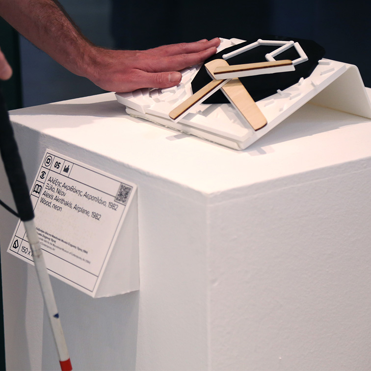 Απτικό βοήθημα για το έργο Αεροπλάνο του Αλέξη Ακριθάκη. Πρόκειται για μια αναπαραγωγή του πρωτότυπου έργου με την τεχνολογία της τρισδιάστατης εκτύπωσης σε λευκό και μαύρο pla πλαστικό, που απεικονίζει απτικά το έργο τέχνης σε κλίμακα 1:10. Η απτική αναπαραγωγή είναι τοποθετημένη με κλίση 30 μοιρών πάνω σε λευκό ξύλινο βάθρο και μπροστά από το βάθρο υπάρχει λεζάντα του έργου με ανάγλυφη μπράιγ και QR κωδικό. Στην εικόνα εμφανίζεται ένα χέρι που διερευνά απτικά το έργο.