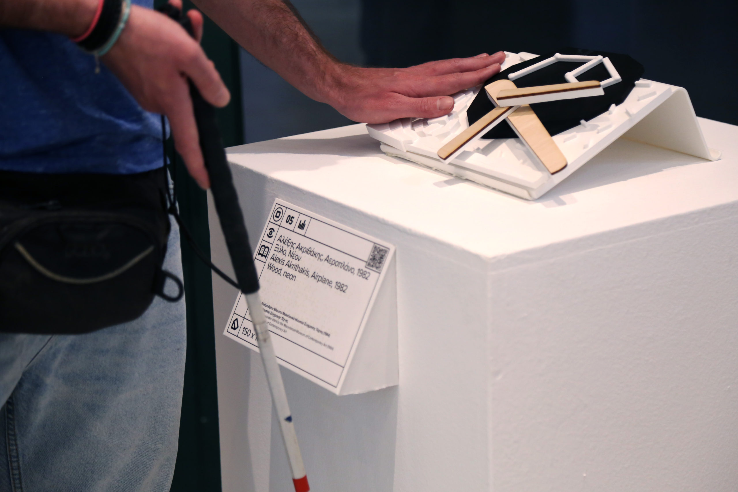 Απτικό βοήθημα για το έργο Αεροπλάνο του Αλέξη Ακριθάκη. Πρόκειται για μια αναπαραγωγή του πρωτότυπου έργου με την τεχνολογία της τρισδιάστατης εκτύπωσης σε λευκό και μαύρο pla πλαστικό, που απεικονίζει απτικά το έργο τέχνης σε κλίμακα 1:10. Η απτική αναπαραγωγή είναι τοποθετημένη με κλίση 30 μοιρών πάνω σε λευκό ξύλινο βάθρο και μπροστά από το βάθρο υπάρχει λεζάντα του έργου με ανάγλυφη μπράιγ και QR κωδικό. Στην εικόνα εμφανίζεται ένα χέρι που διερευνά απτικά το έργο.